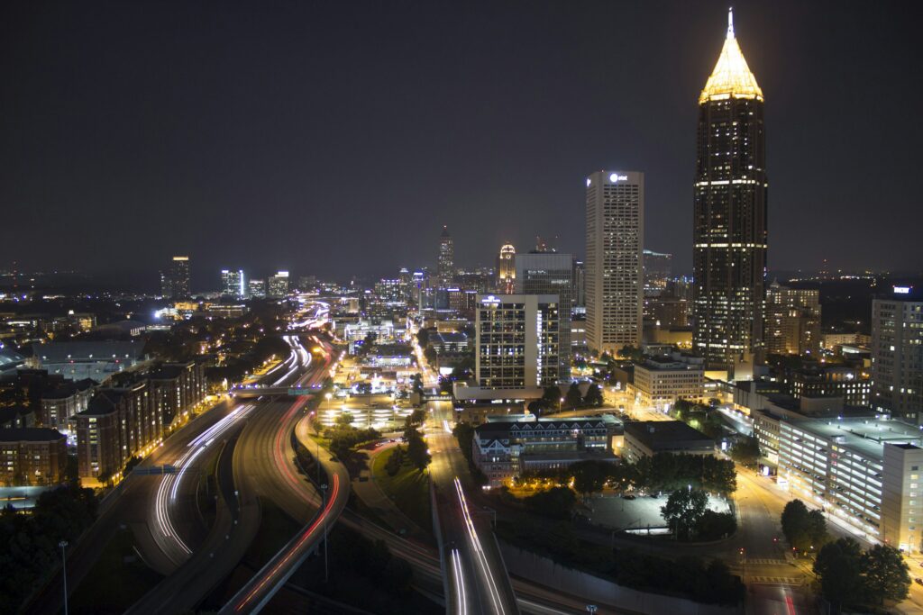 A nighttime aerial view of a cityscape with illuminated skyscrapers and buildings, reminiscent of the advanced IT infrastructure in Contra Costa County. Light trails from vehicles streak through the highways, while the tallest building proudly displays a brightly lit, triangular top.