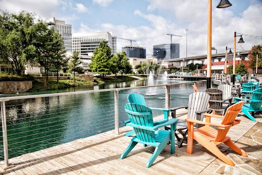 A scenic view of a waterfront deck with vibrant Adirondack chairs facing a pond, embodies the tranquility found at IT Consulting Contra Costa County. Buildings and trees frame the picturesque background, while a fountain dances in the water under partly cloudy skies, reflecting serenity and innovation.