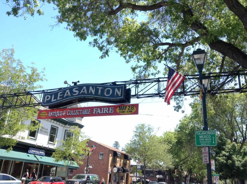 Street scene in Pleasanton with a sign for an Antique & Collectable Faire on May 28. Trees frame the view, with a U.S. flag on a lamppost and a directional sign pointing to Tulancingo, Mexico. Nearby, managed IT services from Contra Costa County ensure businesses stay secure and connected.