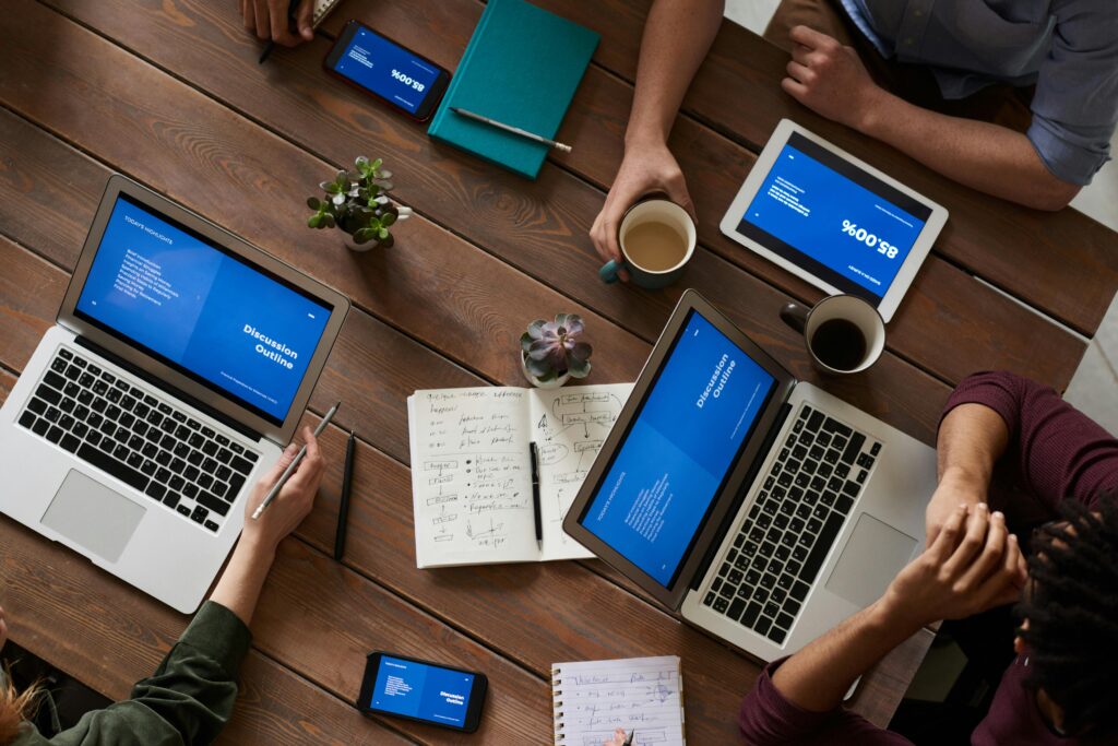 Top view of a wooden table in Contra Costa County with six people using laptops, tablets, and smartphones displaying a presentation. There's a notebook with sketches in the center, alongside coffee cups, pens, and a small plant—a hub for IT support and network security discussions.