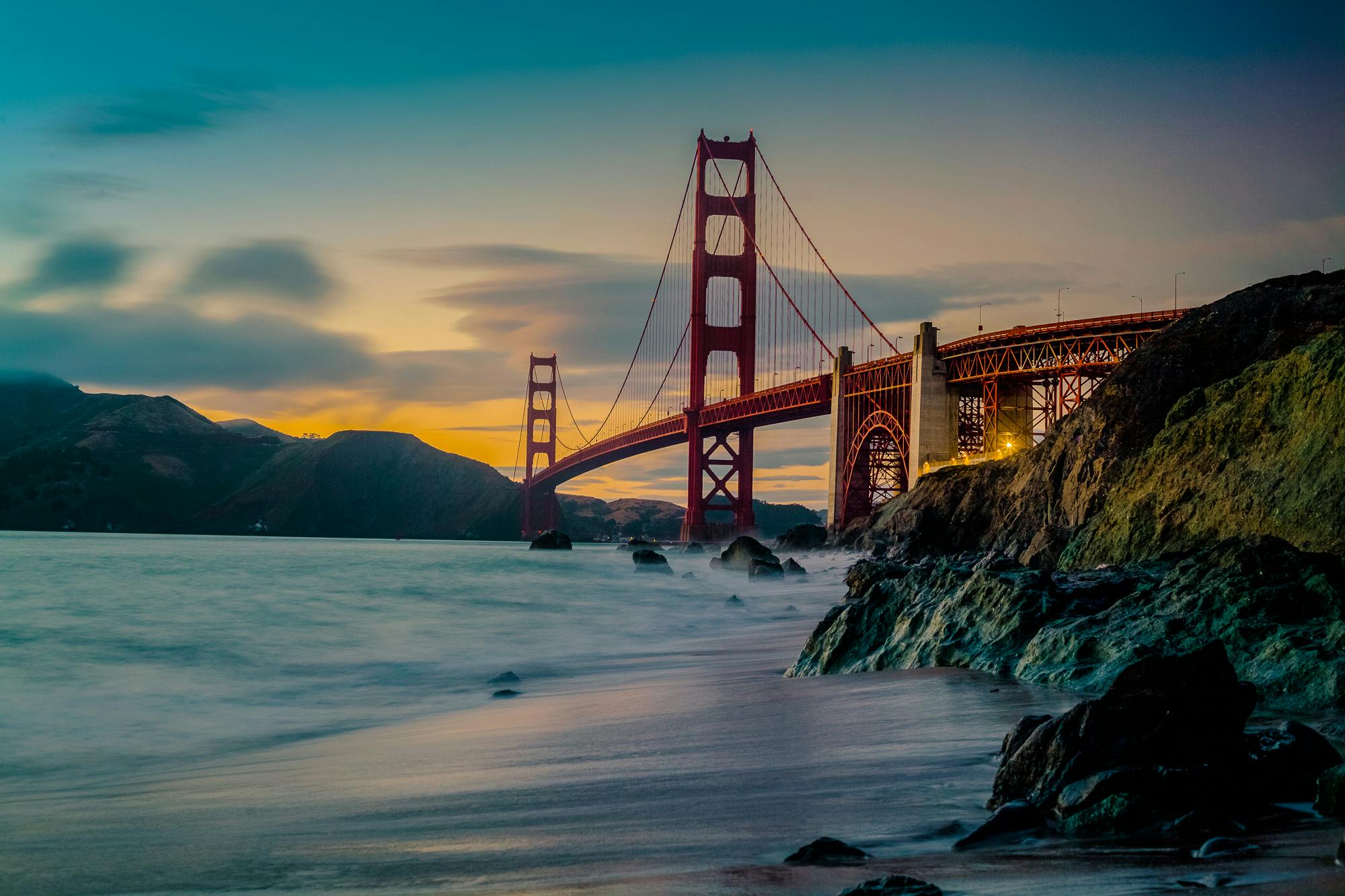 A scenic view of the Golden Gate Bridge at sunset, with the sky lit in shades of orange and blue, reminiscent of the innovative horizons in IT consulting. The bridge spans calm waters, rocky formations visible in the foreground, while distant hills mirror Contra Costa County's managed IT services landscape.