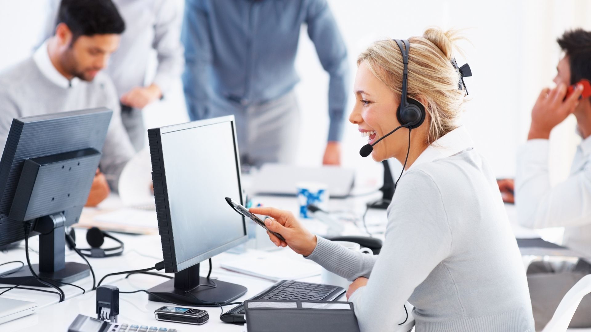 A woman wearing a headset smiles while looking at her smartphone in a modern office, representing IT consulting in Contra Costa County. She sits at a desk with a computer. Two men are in the background, engaged in conversation. The scene is bright and professional.