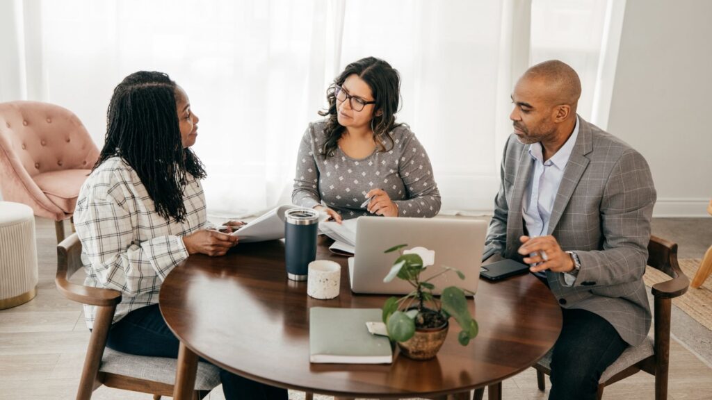 Three people sit around a wooden table in a bright room, engaged in a discussion about IT consulting in Contra Costa County. A laptop, coffee mug, and small plant are on the table. The atmosphere appears collaborative and focused.