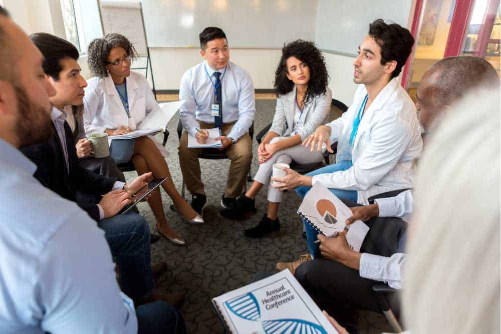 A group of professionals, including doctors and businesspeople, are seated in a circle having a discussion about IT consulting in Contra Costa County. One individual speaks while others listen attentively. Some hold notebooks titled "Annual Healthcare Conference." A flipchart is in the background.