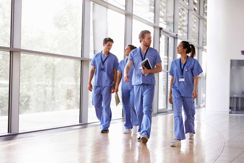 A group of four healthcare professionals in blue scrubs walk together in a hospital hallway with large windows. They are engaged in conversation about IT solutions for Contra Costa County, carrying files and a tablet, and wearing ID badges. Bright natural light fills the space.