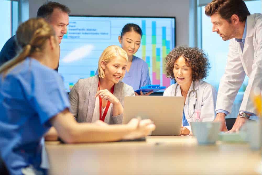 A diverse group of medical professionals gathers around a laptop in Contra Costa County, discussing in a meeting room. A screen displays colorful charts and cybersecurity services insights in the background, indicating a collaborative work environment. Some are dressed in lab coats, others in scrubs.