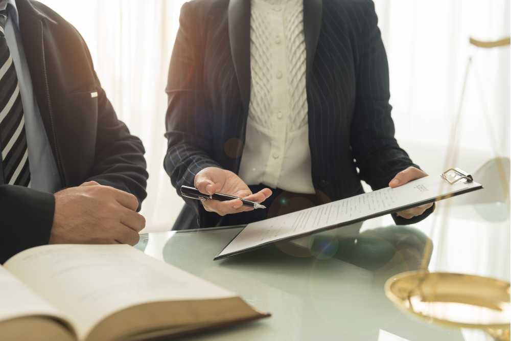 Two professionals in suits are seated at a table. One is holding a clipboard with documents, offering a pen to the other, who has an open book. A balance scale hints at IT support in Contra Costa County, symbolizing the precision and trust needed in tech consultations.