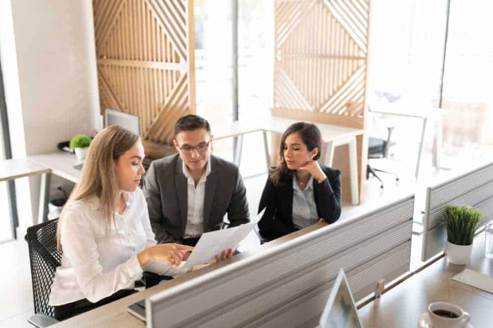 Three people in business attire are seated in a modern office with large windows and wooden accents, reviewing a document. One woman is explaining something related to IT Solutions in Contra Costa County, while the man and another woman listen attentively.