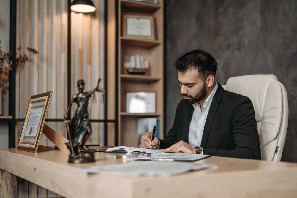 A man in a suit sits at a wooden desk cluttered with paper and a statue of Lady Justice, writing in a book. Shelves with certificates and a decorative ship reflect his IT consulting expertise in Contra Costa County, while the warmly lit room exudes a sense of focus and dedication.