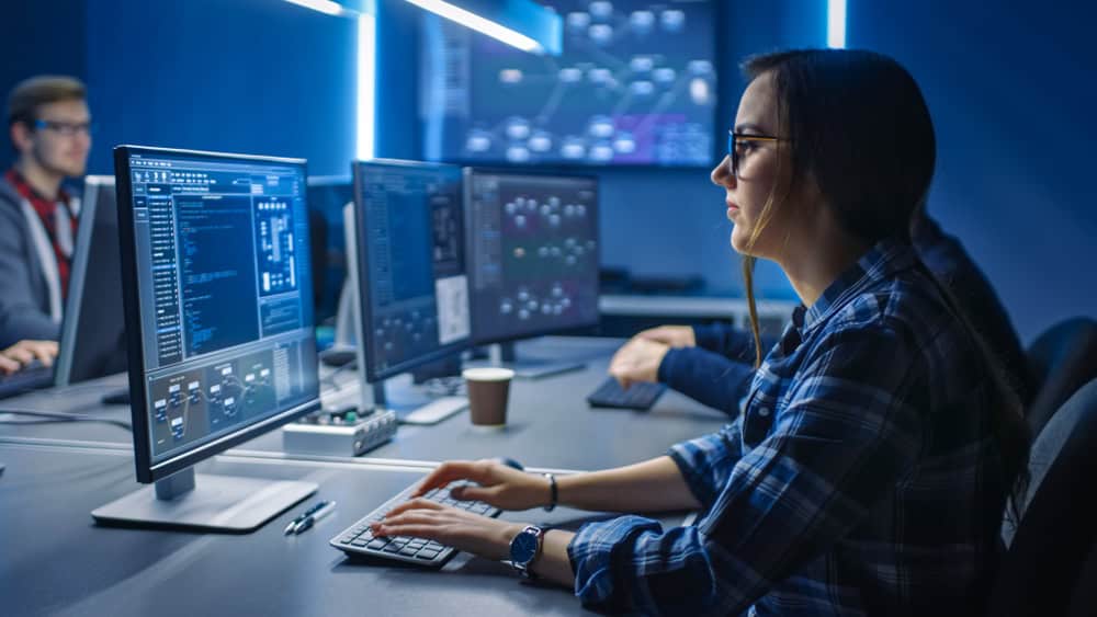 A person wearing glasses and a plaid shirt works at a computer in a dimly lit office, focused on code related to network security in Contra Costa County. Other monitors and people are visible in the background, suggesting an IT consulting or software development environment.