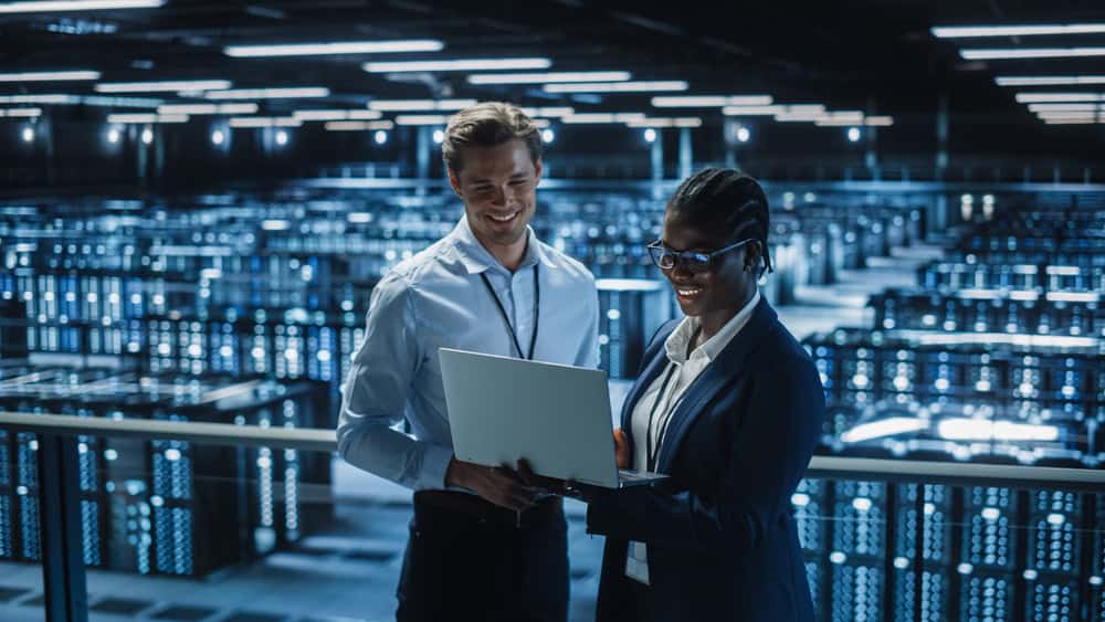 Two people in business attire smile while looking at a laptop in a large, illuminated server room. Rows of servers fill the background, indicating a high-tech or data center environment.