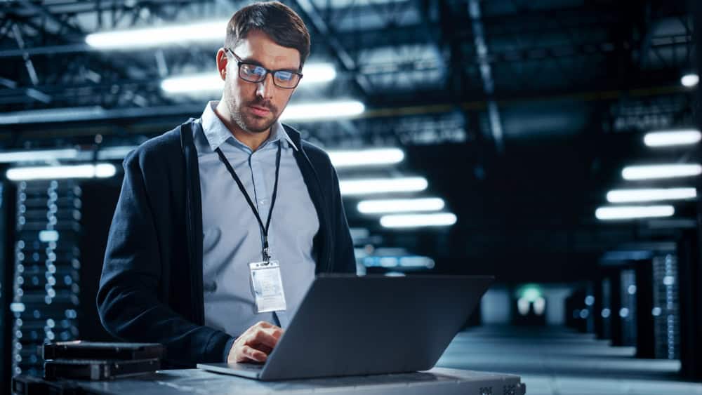 A man wearing glasses and a lanyard works on a laptop in a large, dimly lit data center. Rows of server racks are visible in the background, with bright overhead lights illuminating the space.