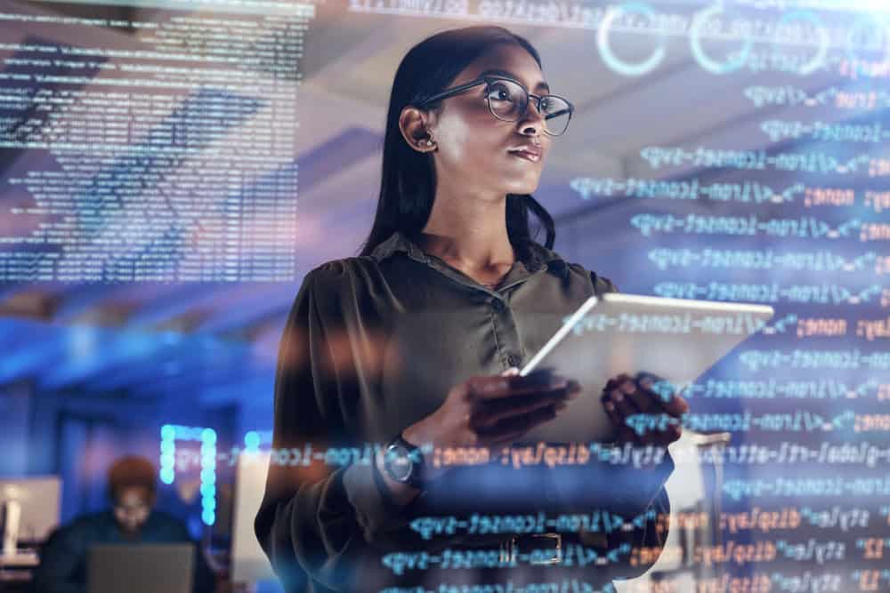 A woman in an office setting holds a tablet and looks intently at a transparent display with code and data visualizations. She wears glasses and a watch, surrounded by a tech environment with servers and multiple screens.