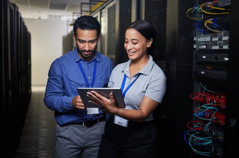 Two IT professionals in an indoor server room, looking at a tablet. They are standing between rows of server racks filled with cables and equipment, wearing ID badges and smiling as they collaborate.