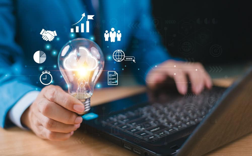 Person in a blue suit holds a glowing lightbulb over a laptop with digital icons representing graphs, people, and a globe overlayed, symbolizing innovation and technology integration.