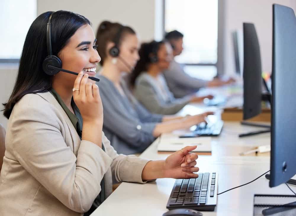 In a bustling call center, employees wearing headsets diligently manage IT support for Contra Costa County. A smiling woman in the foreground efficiently handles inquiries on her computer, while her colleagues engage in similar tasks, ensuring seamless tech assistance for all clients.