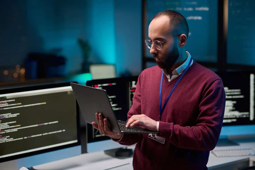 A man with glasses and a beard is typing on a laptop in a dimly lit room with computer screens displaying code in the background. He is wearing a maroon sweater and a lanyard. The atmosphere suggests a tech or programming setting.