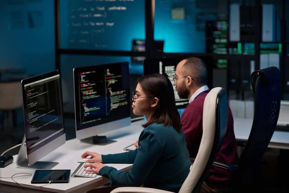 Two people work at desks with computers displaying code. The woman types on a keyboard, while the man looks at his screen. The room is dimly lit, creating a focused atmosphere, with bright monitors as the main light source.