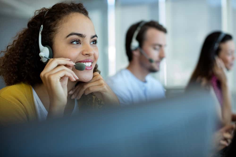 Three customer service representatives wearing headsets are seated and working, focused on providing IT support in Contra Costa County. They appear engaged in their work, sitting in a brightly lit office environment. The foreground shows a blurred computer screen.