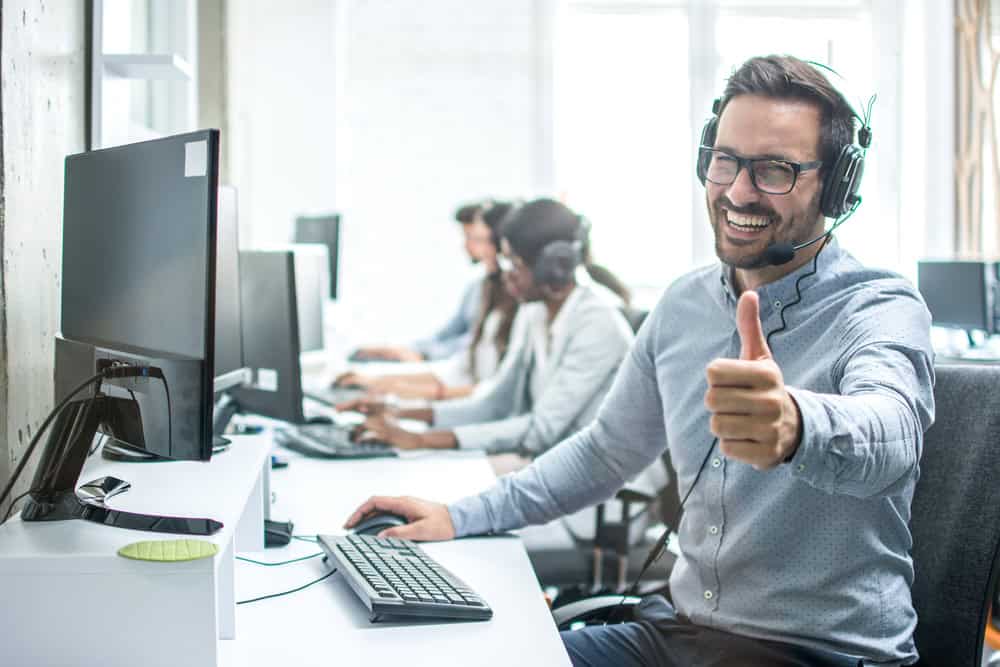 A cheerful man wearing a headset gives a thumbs-up while working at a computer in an office, providing managed IT services in Contra Costa County. In the background, other people are also at desks with headsets, focusing on their screens. The office environment is bright and modern.